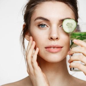 Close up photo of beautiful nude girl smiling looking at camera holding glass of water with cucumber slices over white background. Healthy nutrition. Beauty and skincare.